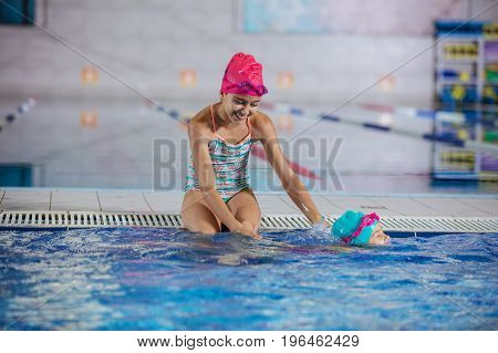 Two sisters in swimming pool: older girl watching younger one swimming along pool nosing