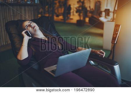 Beautiful caucasian businesswoman is chilling on modern leather massage chair laying relaxed with laptop on her knees and having conversation with her family during lunch time in office chillout