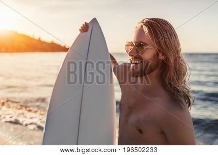 Young handsome man with long hair is standing on beach with white surfboard in hands and smiling.