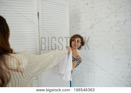 Two girls shopping together: unrecognizable woman giving white dress to attractive young female who is trying on new clothes in fitting-room or cabin. Shopping style fashion and consumerism concept