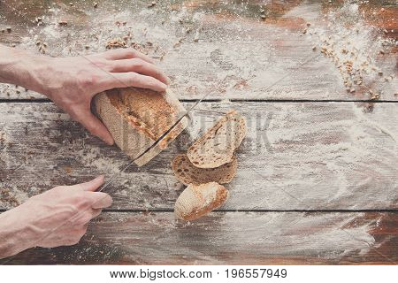 Baking background. Hands of baker cutting bread loaf with knife on rustic wooden table sprinkled with flour.