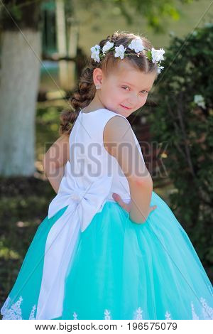 cute girl dressed in blue and white dress with a wreath of artificial flowers on her head child in a festive dress on a nature background half-turn vertical