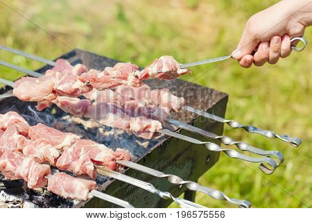 Hand puts skewer with pieces of meat for a shish kebab barbeque on outdoors grill with green blurry background.