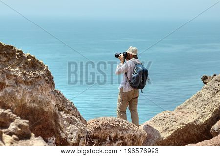 Photographer traveler in a panama on his head and a backpack behind his shoulders stands on the edge of a cliff over the sea Hormuz Island Hormozgan Province southern Iran.