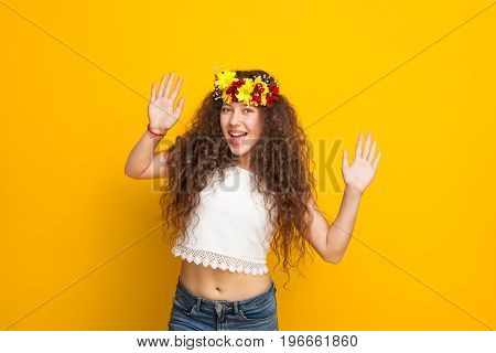 Brunette curly haired girl wearing chaplet white top jeans smiling holding hands up looking at camera.