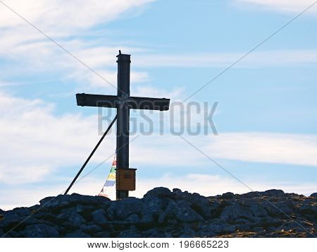 Modest Wooden Cross Raised On Rocky Alpine Mountain Summit . Sharp Rocky Peak. Gentle Clouds  In Blu