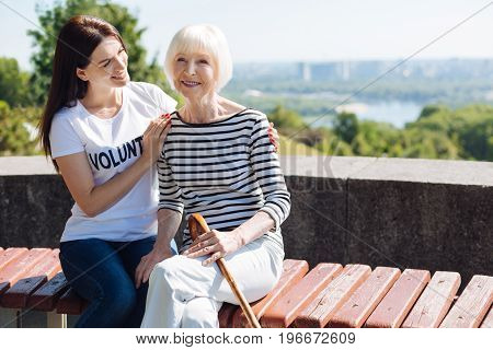 Not alone anymore. Dreamy elegant aged woman feeling thankful while young lady who working as a volunteer keeping her company