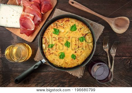 An overhead photo of a Spanish tortilla with potatoes and zucchinis in a tortillera, with jamon, cheese, glasses of white and red wine, a wooden ladle, and two forks, with a place for text