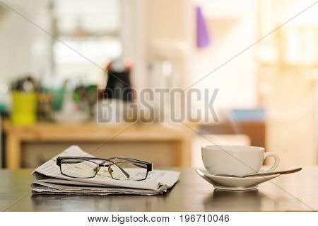 Closeup cup of hot cappuccino coffee with newspaper and spectacles on the wood table with blur kitchen room background-warm tone.