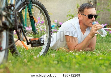 Close up view of man lying on green grass and drinking water from a bottle. Picture of male in white T-shirt and sunglasses with bottle of water lying on green grass with road bicycle beside him in the garden. Selective focus on man. Healthy active lifest