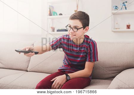 Teenager boy watching television, pointing with remote conroller while sitting on couch in living room at home