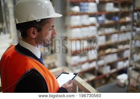 Side view portrait of warehouse supervisor looking down from balcony at tall racks with packed goods holding digital tablet in hand
