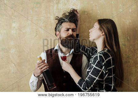 Girlfriend with glass touching messy hair of boyfriend with bottle of wine. Girl or woman and man or hipster with beard on beige wall. Couple in love. Alcohol and convive. Bad habits