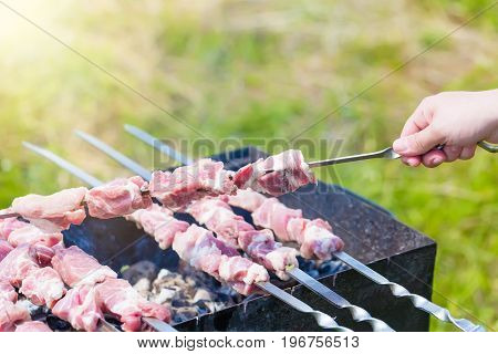 Hand puts skewer with pieces of meat for a shish kebab barbeque on outdoors grill with green blurry background.