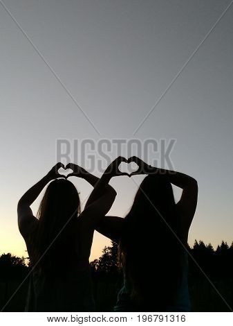 Best friends with interlocking arms and hands making hearts at dusk.