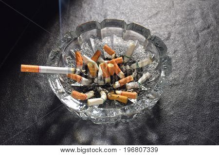 Lit cigarette with smoke lying on an ashtray filled with cigarette butts and ashes