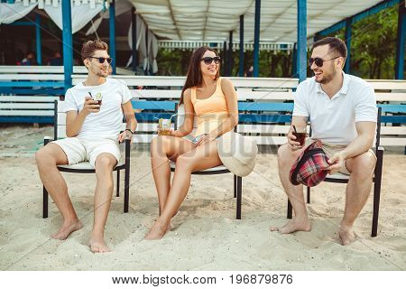 Lifestyle Young people enjoying summer vacation sunbathing drinking at beach bar. Two guys and a young girl sit on the terrace of the summer bar and enjoy the conversation