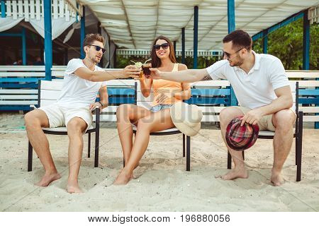 Lifestyle Young people enjoying summer vacation sunbathing drinking at beach bar. Two guys and a young girl sit on the terrace of the summer bar and enjoy the conversation