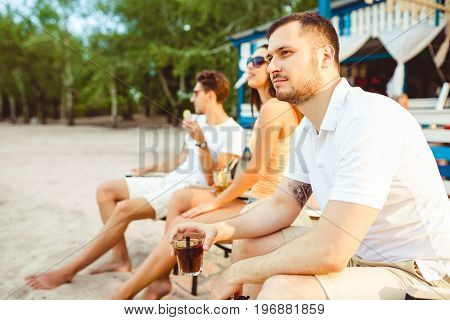 Lifestyle Young people enjoying summer vacation sunbathing drinking at beach bar. Two guys and a young girl sit on the terrace of the summer bar and enjoy the conversation