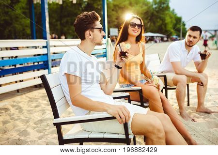 Lifestyle Young people enjoying summer vacation sunbathing drinking at beach bar. Two guys and a young girl sit on the terrace of the summer bar and enjoy the conversation