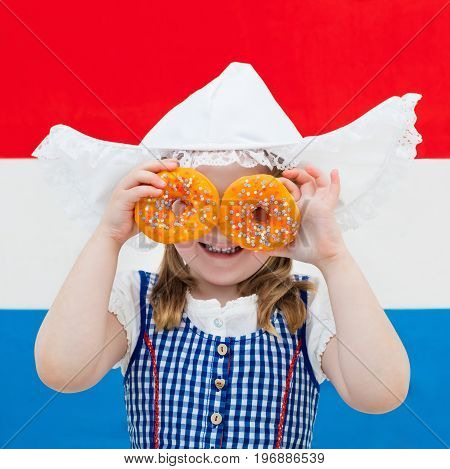 Little Dutch girl wearing traditional national costume dress and hat holding orange donuts at flag of the Netherlands. Child with Oranje souvenirs from Holland celebrating King day. Kid with sweets.
