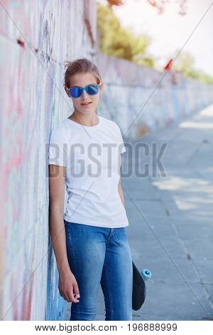 Hipster girl wearing blank white t-shirt, jeans and sunglasses posing against rough street wall, minimalist urban clothing style, mockup for tshirt print store