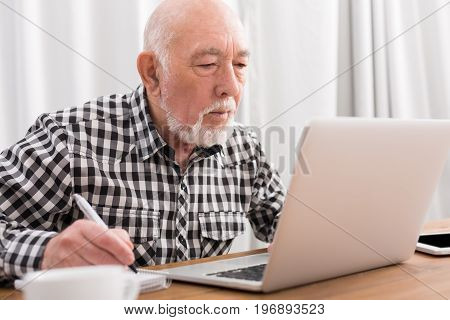 Mature man using laptop and writing in notepad at home desk. Education, recipe concept