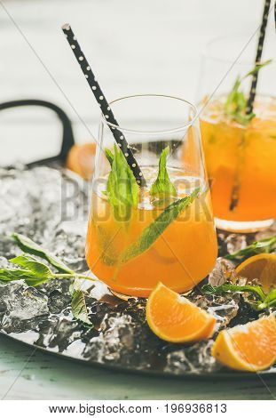 Refreshing cold alcoholic summer citrus cocktail with orange, peppermint and crushed ice in stemless glasses on dark tray over light wooden background, selective focus