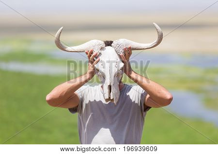 Man holding a white wildebeest skull wearing it like a mask in nature on african wildlife safari, Amboseli national park, Kenya.