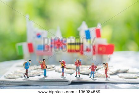 Tiny miniature model of travelers or backpackers are standing on a passport and a pile of coins with international flags background using as traveling journey travel to destination background concept.