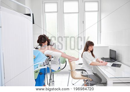 Young nurse writing sitting at the desk while professional gynecologist examining her patient on a gynecological chair hospital clinic femininity healthcare medicine vitality concept.