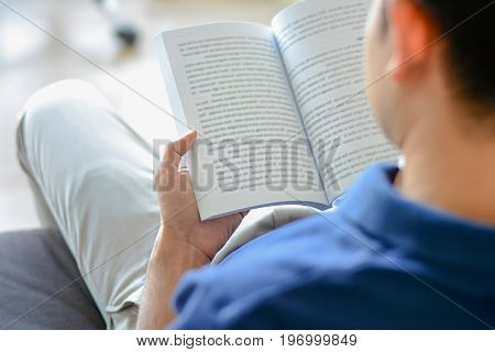 Young man reading book while sitting on the couch over shoulder view with soft focus