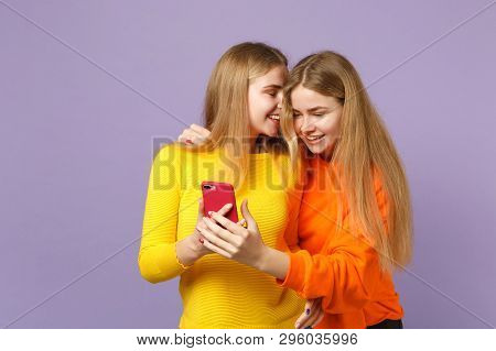 Two Cheerful Young Blonde Twins Sisters Girls In Vivid Clothes Using Mobile Phone, Typing Sms Messag