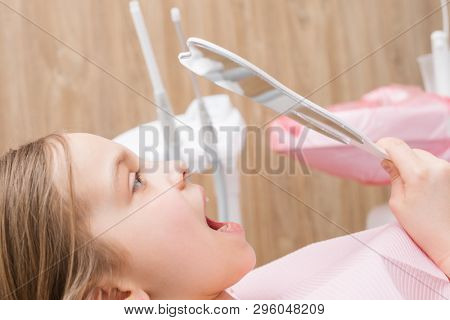 Portrait of elementary age girl sitting in a dental chair inspecting repaired teeth looking thru tooth-shaped mirror in pediatric dental clinic