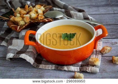 Pumpkin Carrot Cream Soup In An Orange Plate With Crackers
 On A Checked Tablecloth With Wooden Back