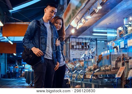Young Asian Couple Looking For Something Special At Traditional Chainese Market. They Are Pensive. A