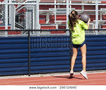 A High School Teenage Girl Is Thowing A Medicine Ball Over Her Head Backwards On A Track In Front Of
