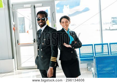 Smiling African American Pilot In Sunglasses And Stewardess With Crossed Arms Standing Together In A