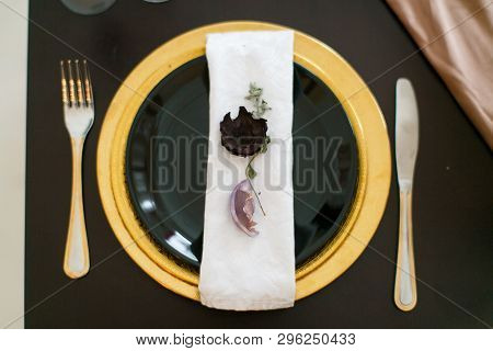 On A Dark Table There Is A Yellow Plate, On It Is A Black Plate With A Napkin, On Which Dried Petals