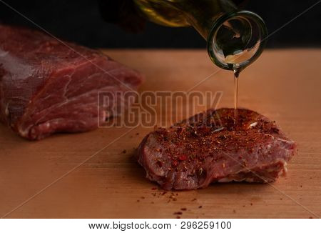 Hand in black glove pouring oil on beef steak with spices on a wooden board with a black background