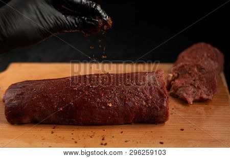 Hand In Black Glove Sprinkling Spice On Beef Steak On A Wooden Board With A Black Background
