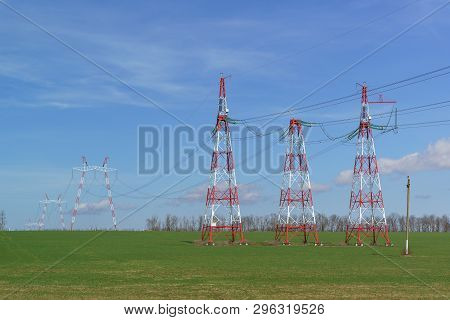 Supports Power Lines Are In The Field. Red And White Against A Bright Blue Sky. Solar Panels For Nig