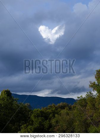 Heart In Cloudy Sky. Blue White Clouds With White Heart. Trees And Mountains In The Background. Lake