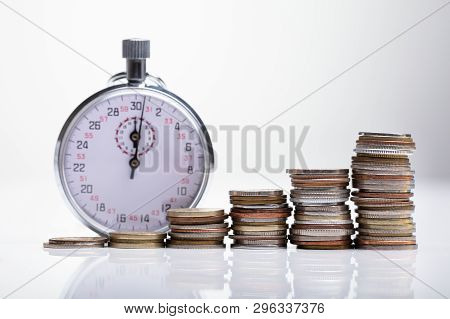 Close-up Of Stopwatch With Increasing Stacked Coins On Reflective Desk