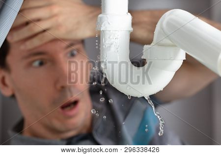 Shocked Young Man Looking At Sink Pipe Leakage In Kitchen