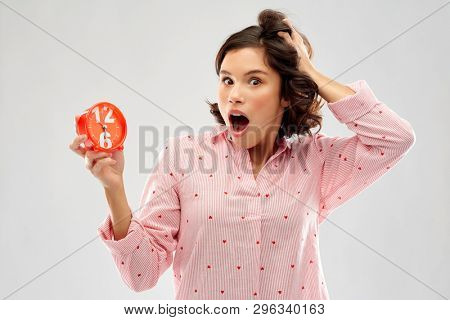 time, emotions and bedtime concept - shocked young woman in pajama with alarm clock showing half past six over grey background