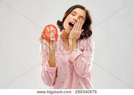 people and bedtime concept - sleepy young woman in pajama with alarm clock showing half past six yawning over grey background