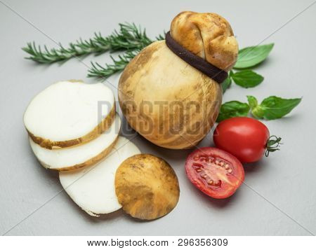 Traditional Italian smoked Scamorza cheese with herbs and tomatoes on gray background.