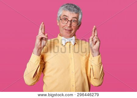 Studio Shot Of White Haired Senior Raises Hands Up And Crosses Fingers. Elderly Man Wears Yellow Shi