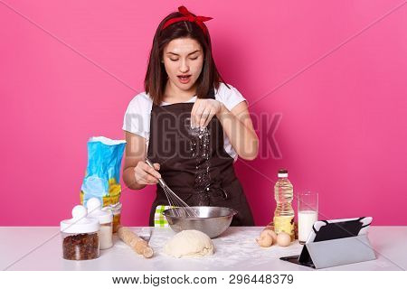 Busy Creative Young Woman Poses In White T Shirt, Red Headband And Brown Apron, Adding Pinch Of Flou